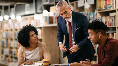 Two students of color talking with a white professor in a library