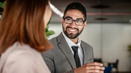 A man in a grey suit chatting with a woman in a pink blouse at work.