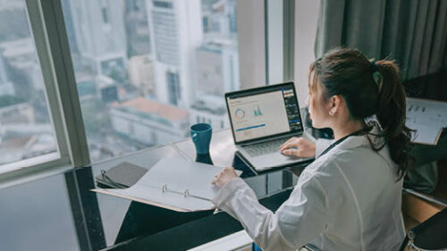 A woman examining a binder of information at a desk with data graphs on a laptop next to her.