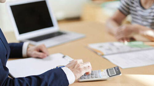 two business professionals sitting at a table with papers, a laptop and calculators scattered on top.