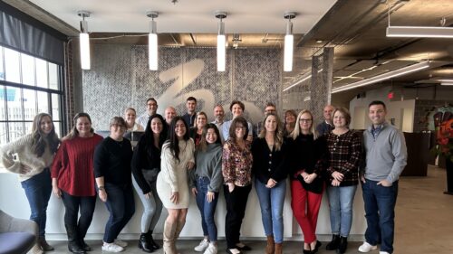 Holmes Murphy Kansas City employees stand in business casual clothes together in front of the white desk that welcomes visitors to Holmes Murphy in Kansas City