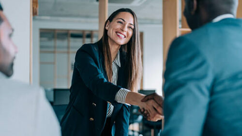 A smiling woman wearing a blazer shaking hands with a man in a suit.