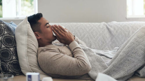 A man laying on a couch while blowing his nose with a tissue.